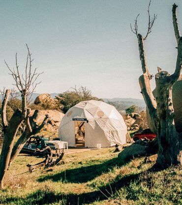 A white geodesic glamping dome with an open entrance, situated in a rugged, sunlit landscape. The dome, bearing a small sign above the door that reads 'THE CROWN', sits on a wooden deck. The scene is framed by two large, bare, dead tree trunks in the foreground. The surrounding area features sparse green grass, large boulders, and a red vehicle partially visible behind the dome, with rolling mountains in the distance under a clear blue sky.