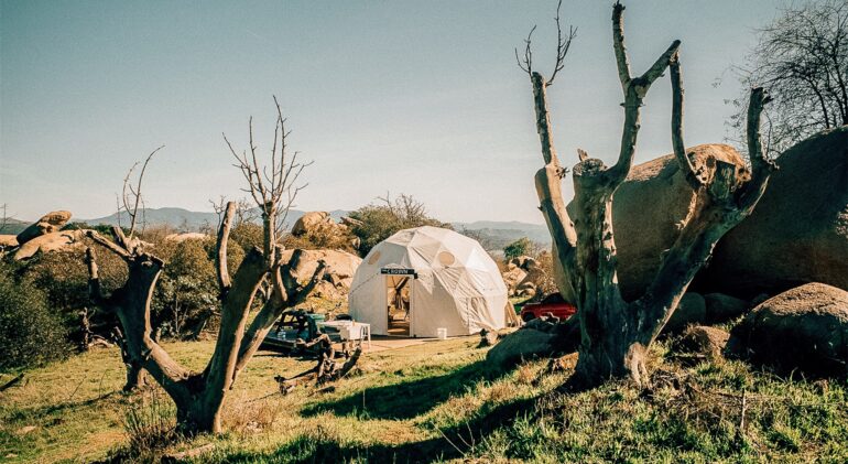 A white geodesic glamping dome with an open entrance, situated in a rugged, sunlit landscape. The dome, bearing a small sign above the door that reads 'THE CROWN', sits on a wooden deck. The scene is framed by two large, bare, dead tree trunks in the foreground. The surrounding area features sparse green grass, large boulders, and a red vehicle partially visible behind the dome, with rolling mountains in the distance under a clear blue sky.
