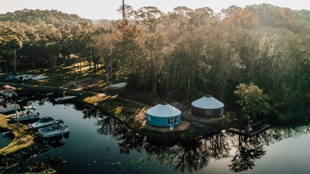 An elevated aerial photograph captures two large, round yurts with light-colored roofs, one teal and one reddish-brown, nestled within a dense forest of trees and vegetation by a calm river at sunrise. A collection of diverse boats is docked on the left bank, and shadows are cast from the trees over a dirt clearing and onto the water, which has a distinct, dark blue-green hue. In the background, there is a clearing with parked cars. The rising sun, located above the right horizon, bathes the scene in warm, hazy light.