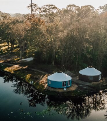 An elevated aerial photograph captures two large, round yurts with light-colored roofs, one teal and one reddish-brown, nestled within a dense forest of trees and vegetation by a calm river at sunrise. A collection of diverse boats is docked on the left bank, and shadows are cast from the trees over a dirt clearing and onto the water, which has a distinct, dark blue-green hue. In the background, there is a clearing with parked cars. The rising sun, located above the right horizon, bathes the scene in warm, hazy light.