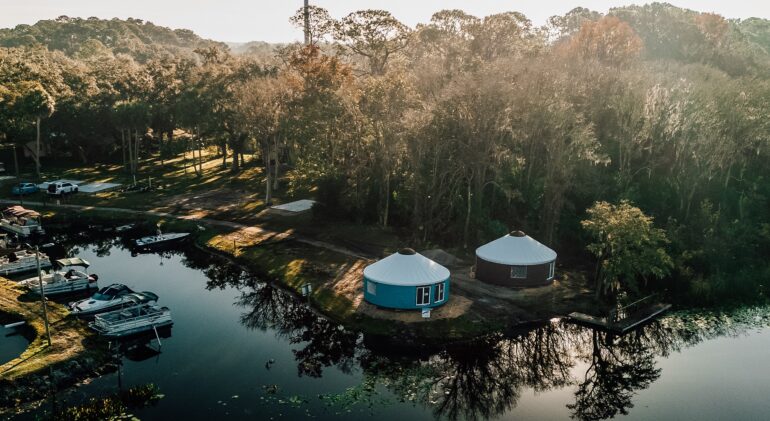 An elevated aerial photograph captures two large, round yurts with light-colored roofs, one teal and one reddish-brown, nestled within a dense forest of trees and vegetation by a calm river at sunrise. A collection of diverse boats is docked on the left bank, and shadows are cast from the trees over a dirt clearing and onto the water, which has a distinct, dark blue-green hue. In the background, there is a clearing with parked cars. The rising sun, located above the right horizon, bathes the scene in warm, hazy light.