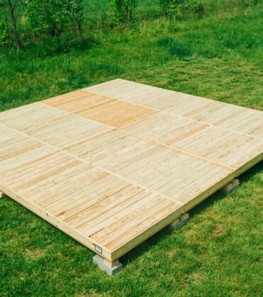 A large, square modular wooden deck platform made of light-colored wood panels with a checkerboard pattern is assembled on a green grass field. The platform is leveled and raised slightly on small concrete supports, with a line of trees in the background.