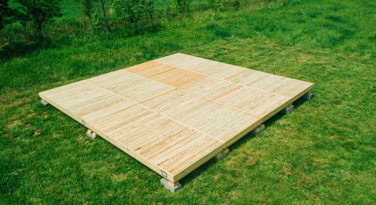 A large, square modular wooden deck platform made of light-colored wood panels with a checkerboard pattern is assembled on a green grass field. The platform is leveled and raised slightly on small concrete supports, with a line of trees in the background.