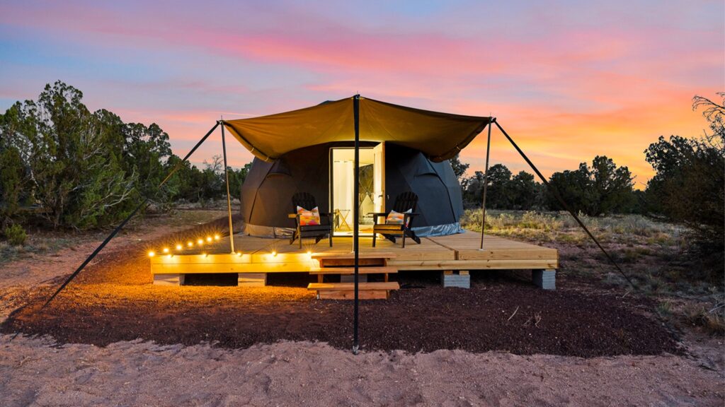 A black glamping dome tent on a raised wooden deck in a desert landscape at sunset. A tan awning stretches over two black Adirondack chairs with colorful pillows, while warm string lights illuminate the edge of the deck. The open door reveals a brightly lit interior, contrasting beautifully against a vibrant pink, orange, and blue sky.