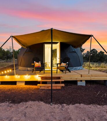 A black glamping dome tent on a raised wooden deck in a desert landscape at sunset. A tan awning stretches over two black Adirondack chairs with colorful pillows, while warm string lights illuminate the edge of the deck. The open door reveals a brightly lit interior, contrasting beautifully against a vibrant pink, orange, and blue sky.