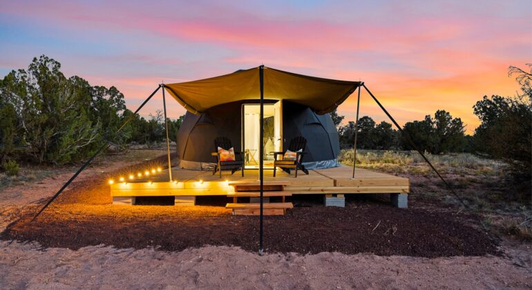 A black glamping dome tent on a raised wooden deck in a desert landscape at sunset. A tan awning stretches over two black Adirondack chairs with colorful pillows, while warm string lights illuminate the edge of the deck. The open door reveals a brightly lit interior, contrasting beautifully against a vibrant pink, orange, and blue sky.
