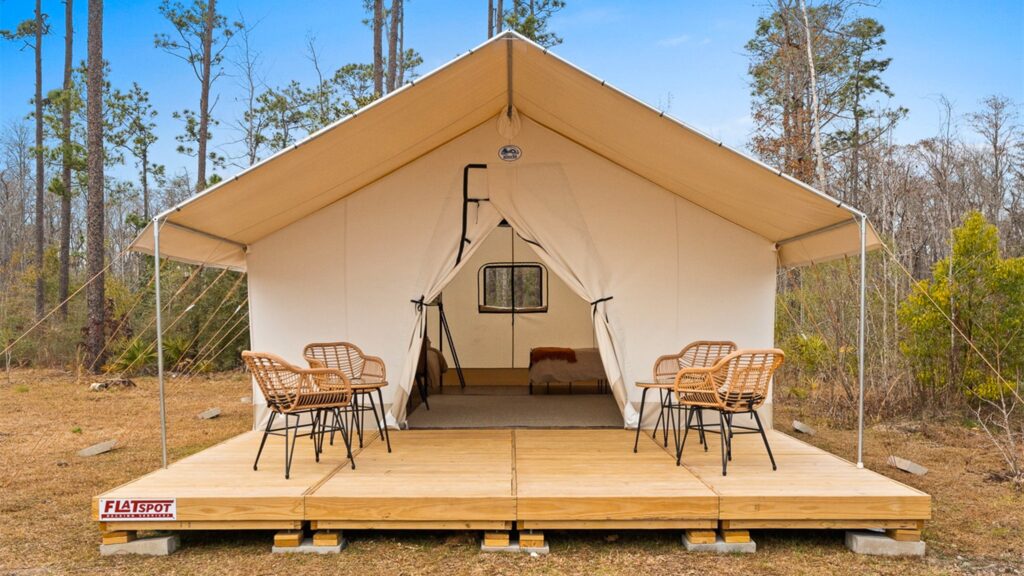 A large, canvas safari-style glamping tent pitched on an elevated wooden deck in a wooded area. The front flaps of the tent are tied open, revealing a bed inside. On the deck outside the tent, four wicker chairs are arranged around two small tables.