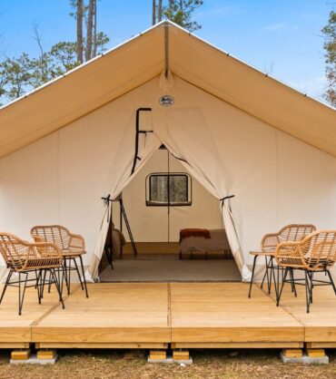 A large, canvas safari-style glamping tent pitched on an elevated wooden deck in a wooded area. The front flaps of the tent are tied open, revealing a bed inside. On the deck outside the tent, four wicker chairs are arranged around two small tables.