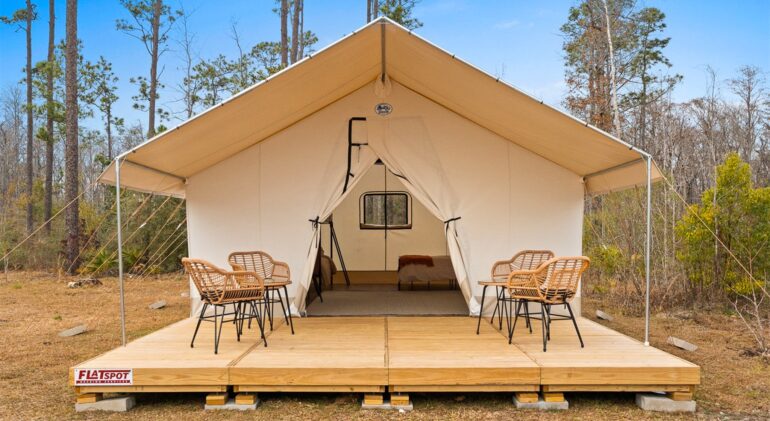 A large, canvas safari-style glamping tent pitched on an elevated wooden deck in a wooded area. The front flaps of the tent are tied open, revealing a bed inside. On the deck outside the tent, four wicker chairs are arranged around two small tables.